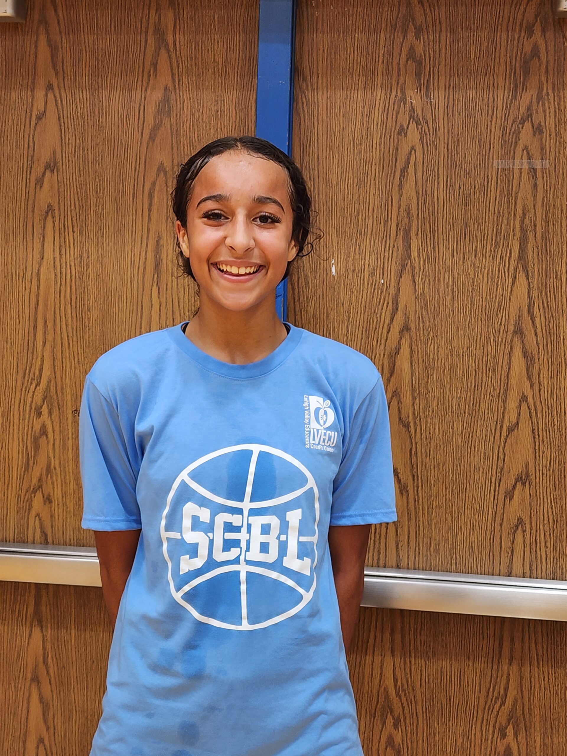 A smiling SCBL youth basketball player stands in a blue team shirt during the Highlight Reel Photo Gallery, framed by wood-paneled gym doors behind.
