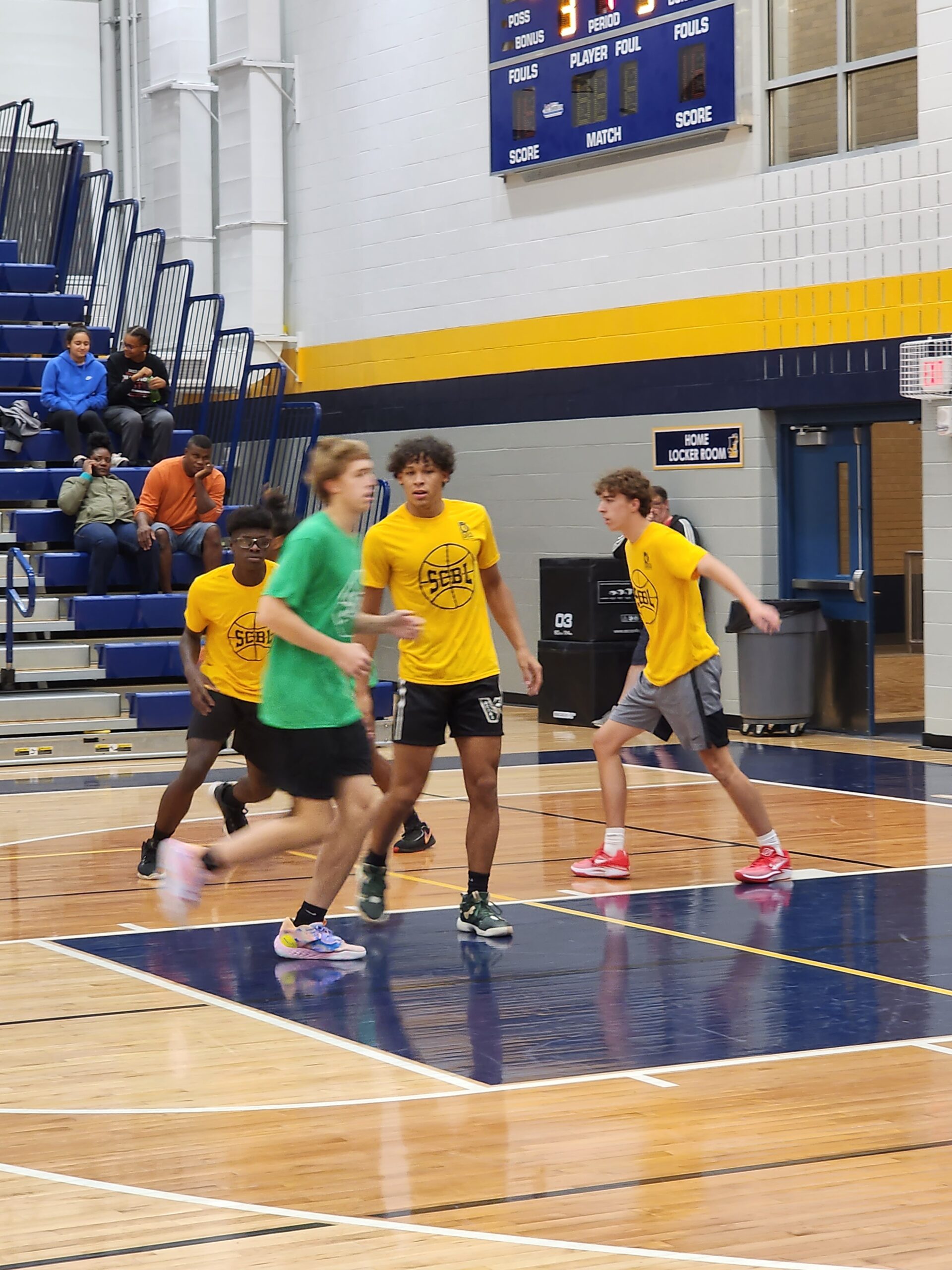 During an SCBL basketball game in the gym, players in yellow and green jerseys move across the court as spectators watch from the bleachers.