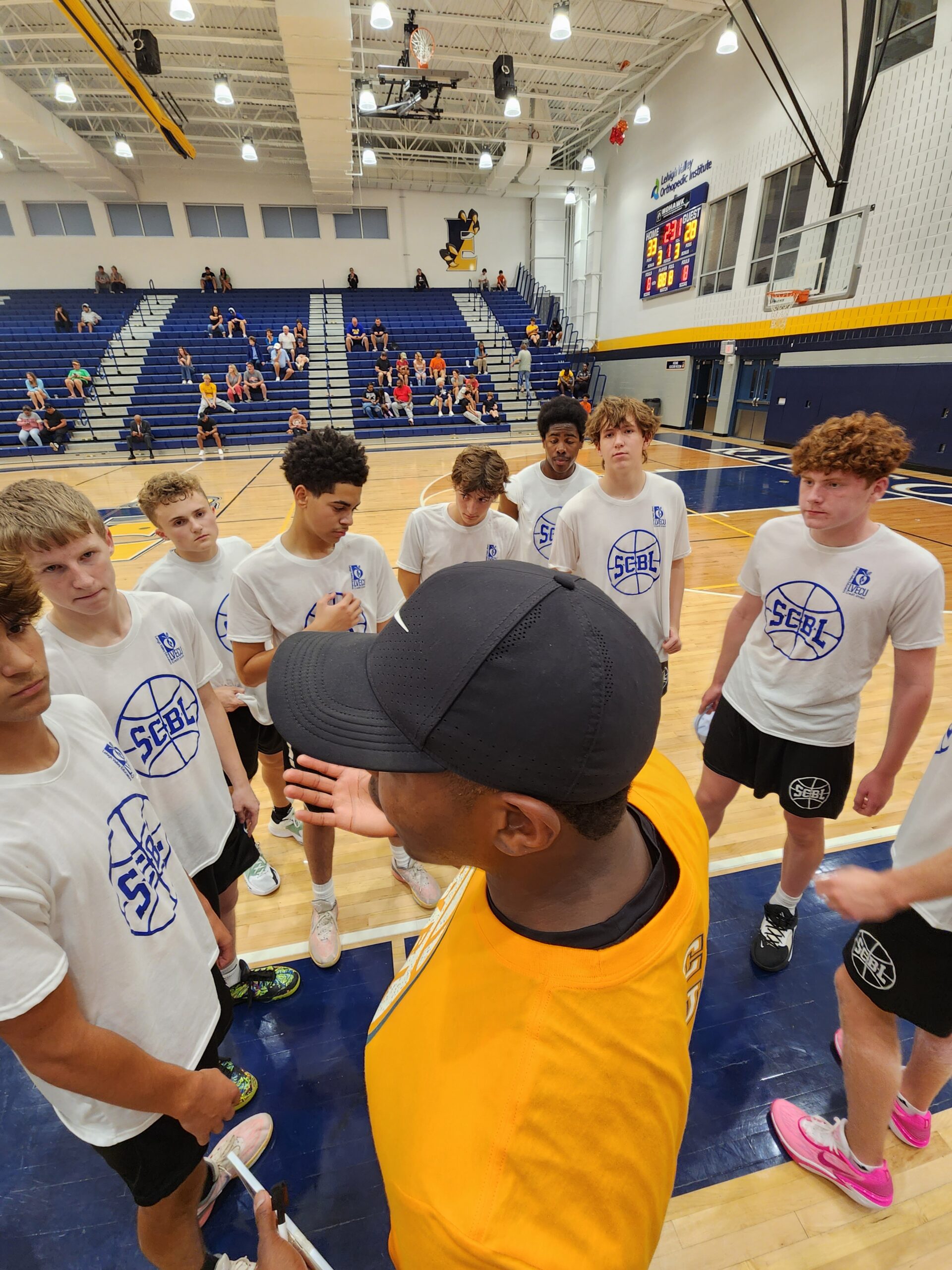 During an SCBL basketball huddle, players in white shirts gather around a coach on an indoor court while fans sit in the bleachers.