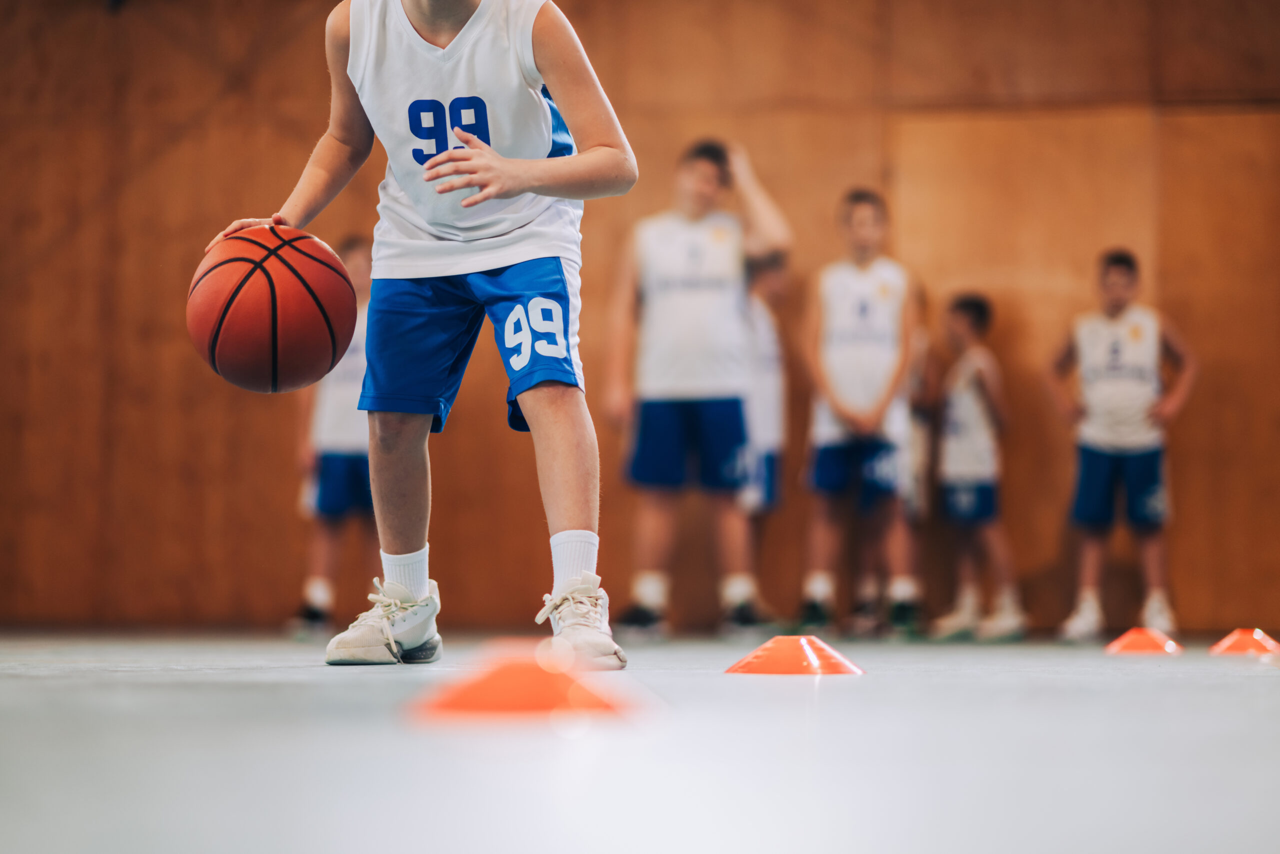 During SCBL Programs, a young player dribbles through cone drills in the gym while teammates wait nearby and watch the training session.