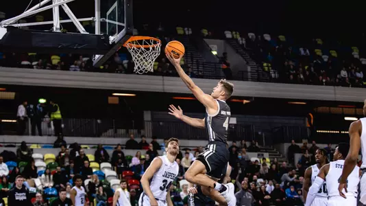 During an SCBL basketball game, Chris Mann drives to the rim for a one-handed dunk as defenders and spectators watch from the lane and stands.