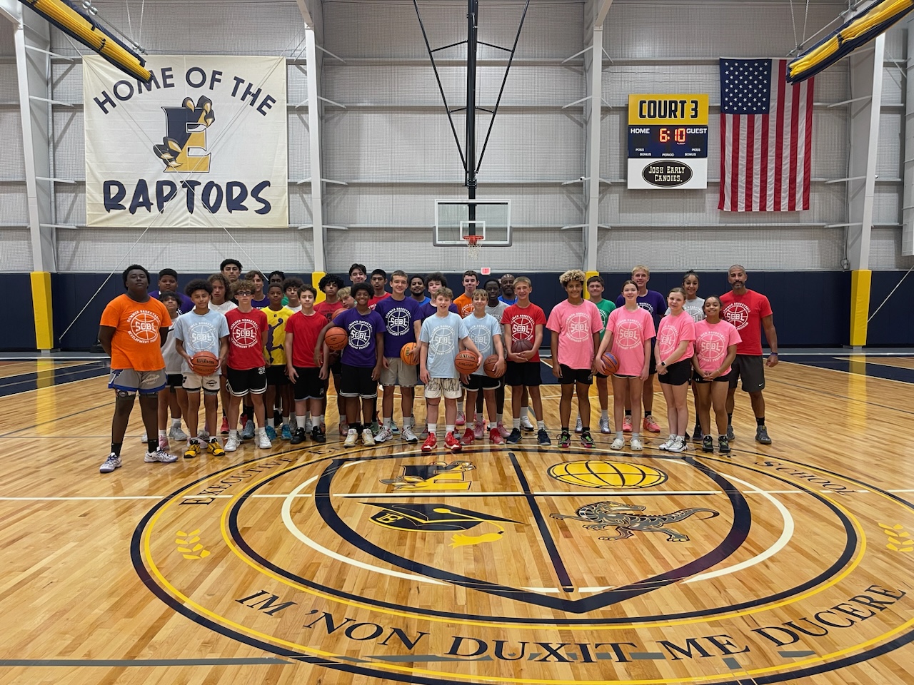On Court 3 at SCBL, a large group of players and coaches poses for a gallery moment beneath the Raptors banner and scoreboard while holding basketballs together.