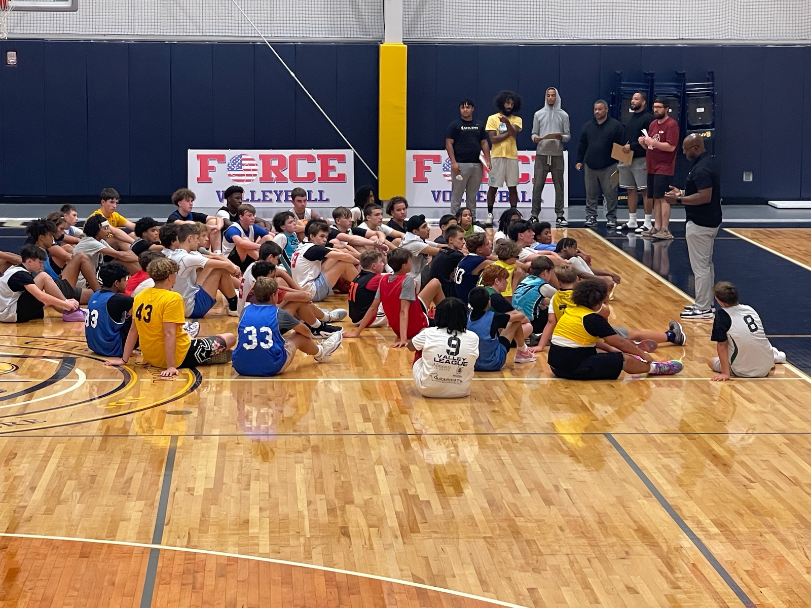 At SCBL Gallery, youth basketball players sit on the court for a team huddle while coaches speak in a gym with a FORCE VOLLEYBALL sign behind them during practice.
