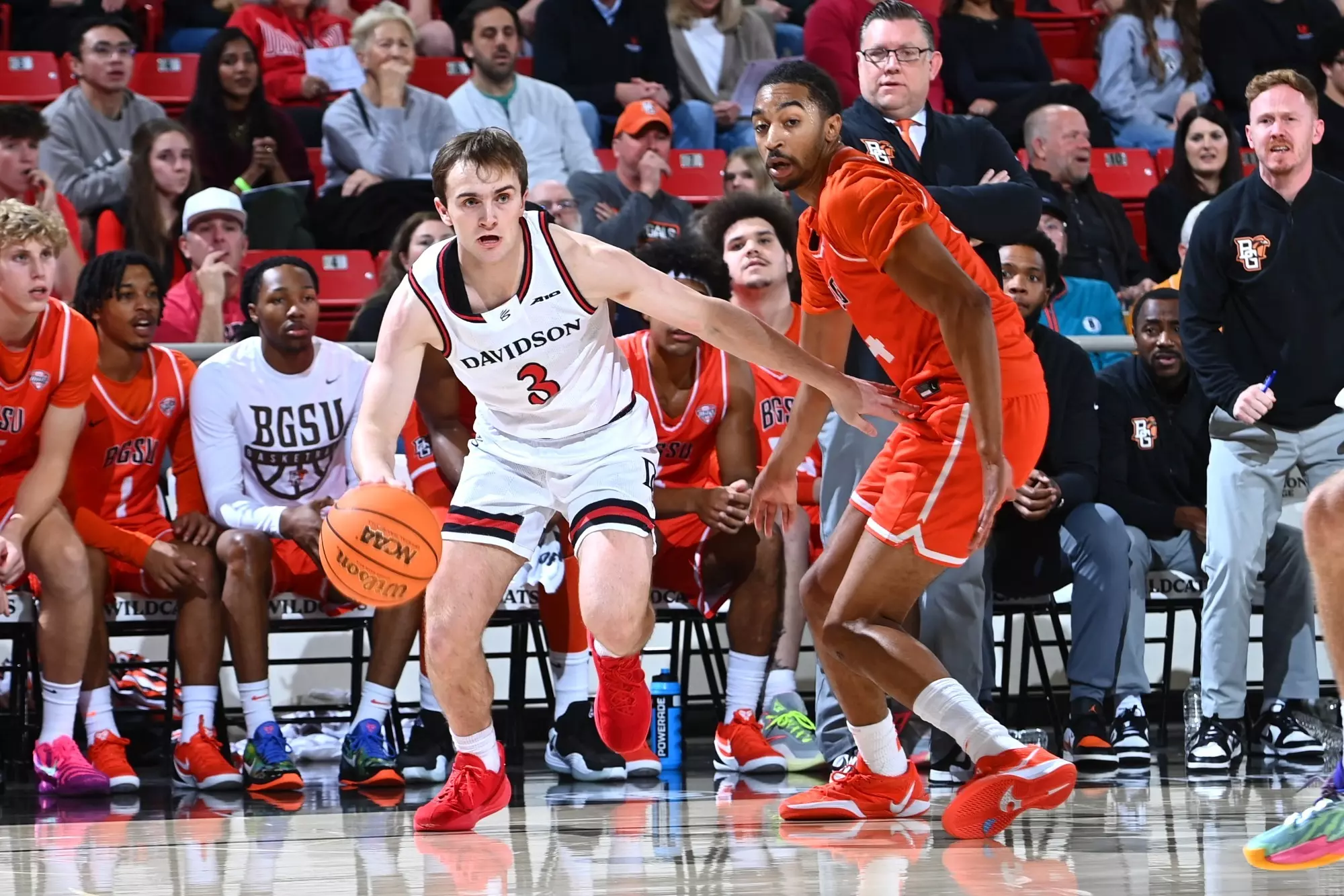 Nick Coval dribbles for Davidson against a Bowling Green defender during a fast-paced college basketball game, with SCBL benches and fans behind.