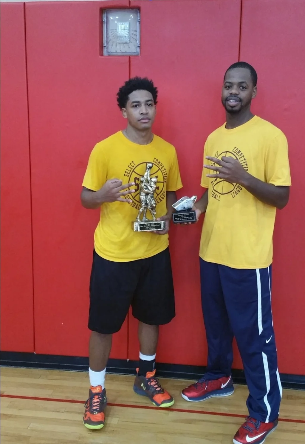 Two SCBL players in yellow shirts pose with championship trophies and hand signs in front of a red gym wall at the court after a game.