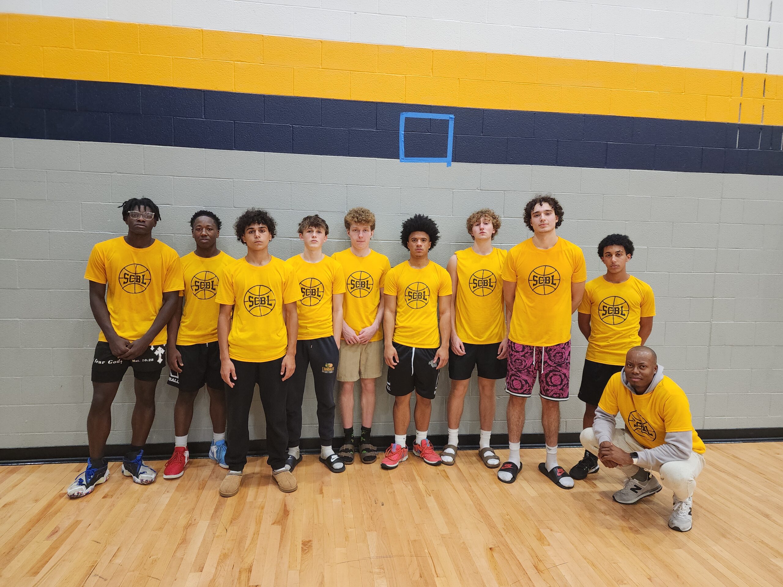 A group of SCBL boys basketball players and a coach stand together in matching yellow shirts during team programs inside a gym.
