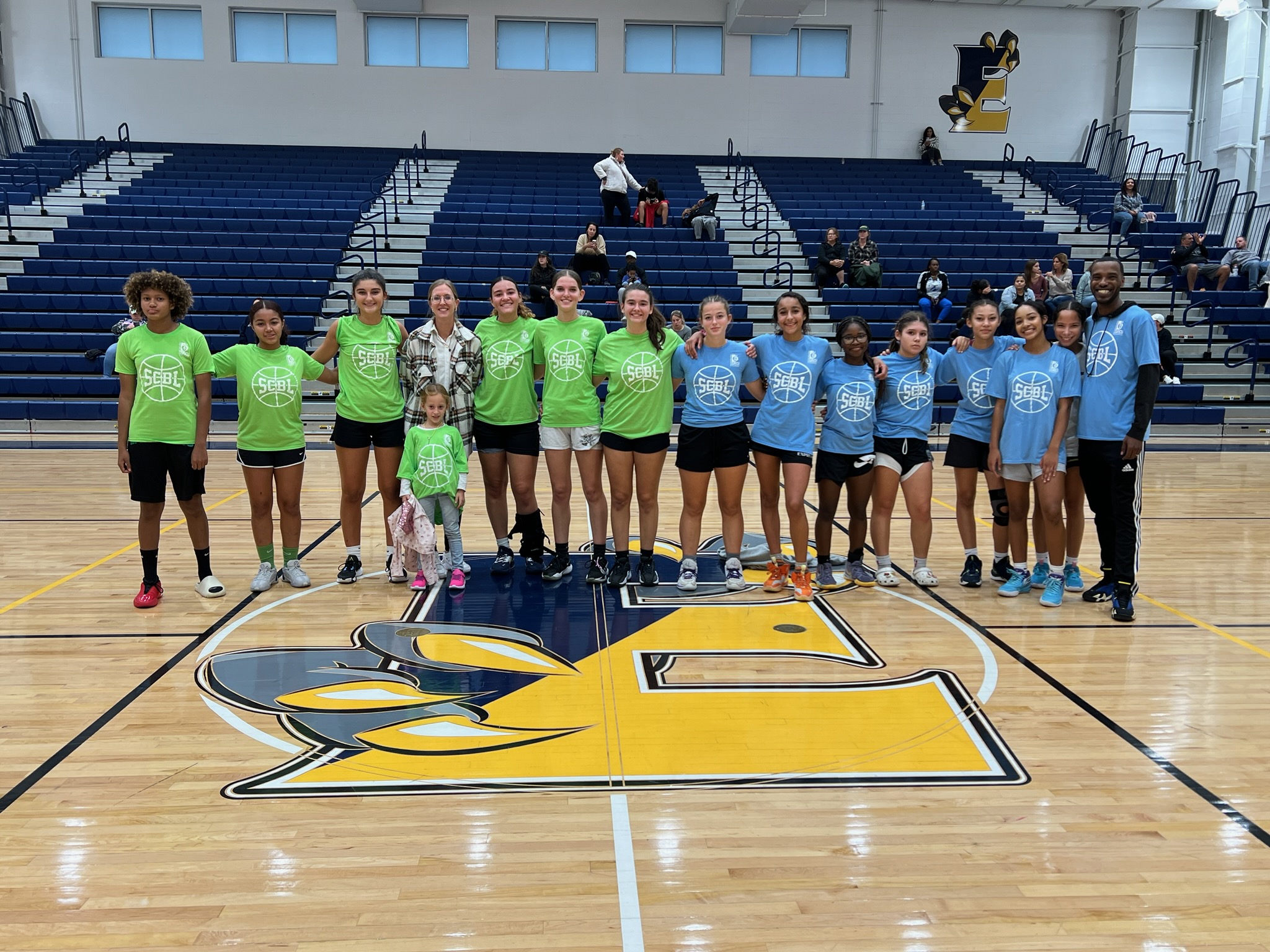 SCBL players and coaches line up on the gym floor after a competition, wearing green and blue shirts at center court in a school gym.