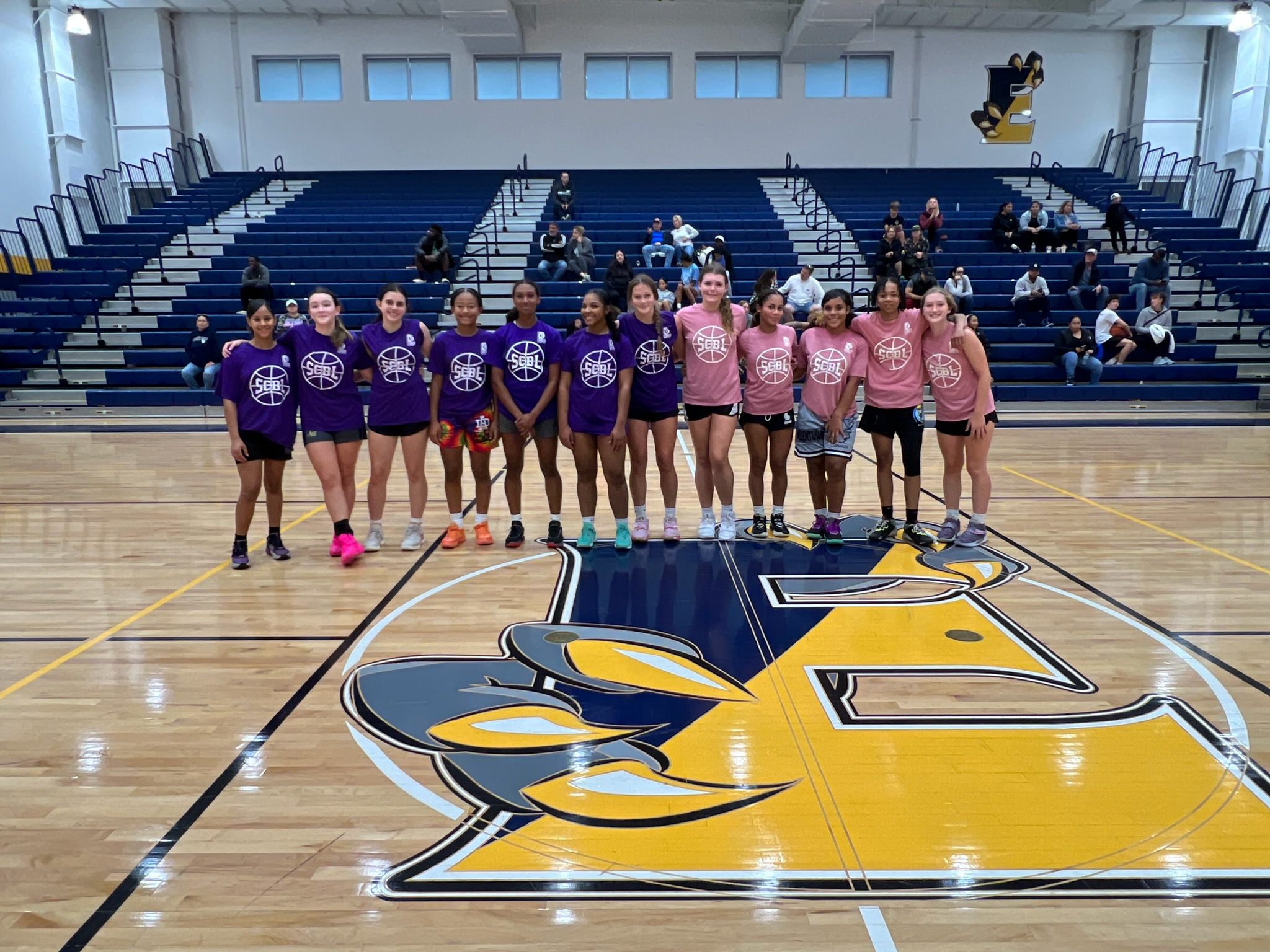 A group of girls in SCBL shirts stands together on the basketball court at a girls basketball programs event inside a school gym for team photos.