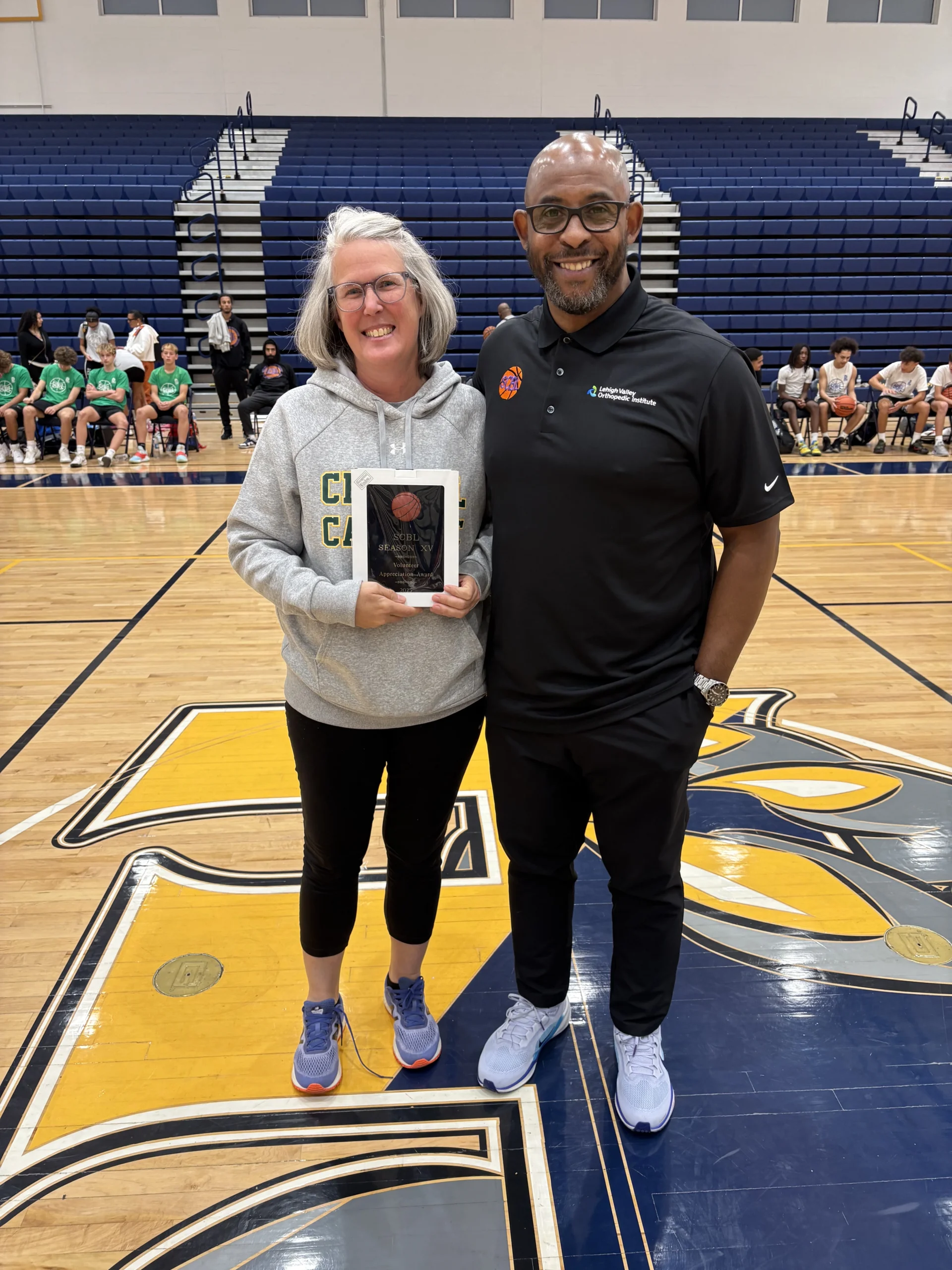 At an SCBL youth basketball event, a woman receives a volunteer appreciation award beside a coach in a Lehigh Valley Orthopedic Institute polo.