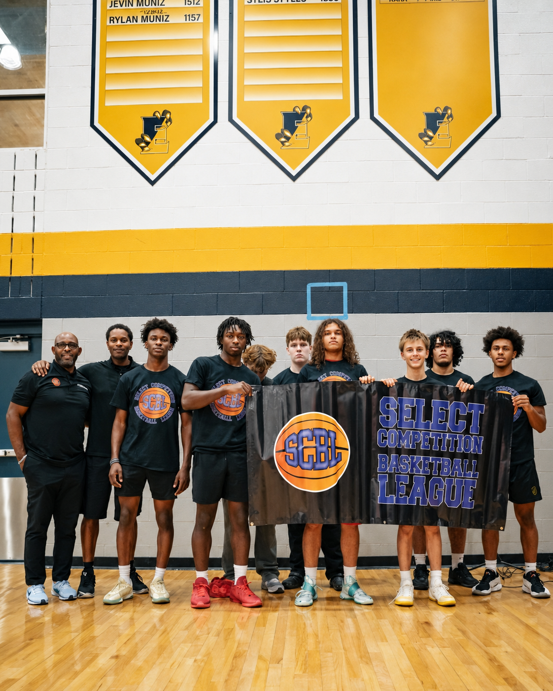 SCBL team members and coaches stand together in black uniforms holding a Select Competition Basketball League banner inside a gym.